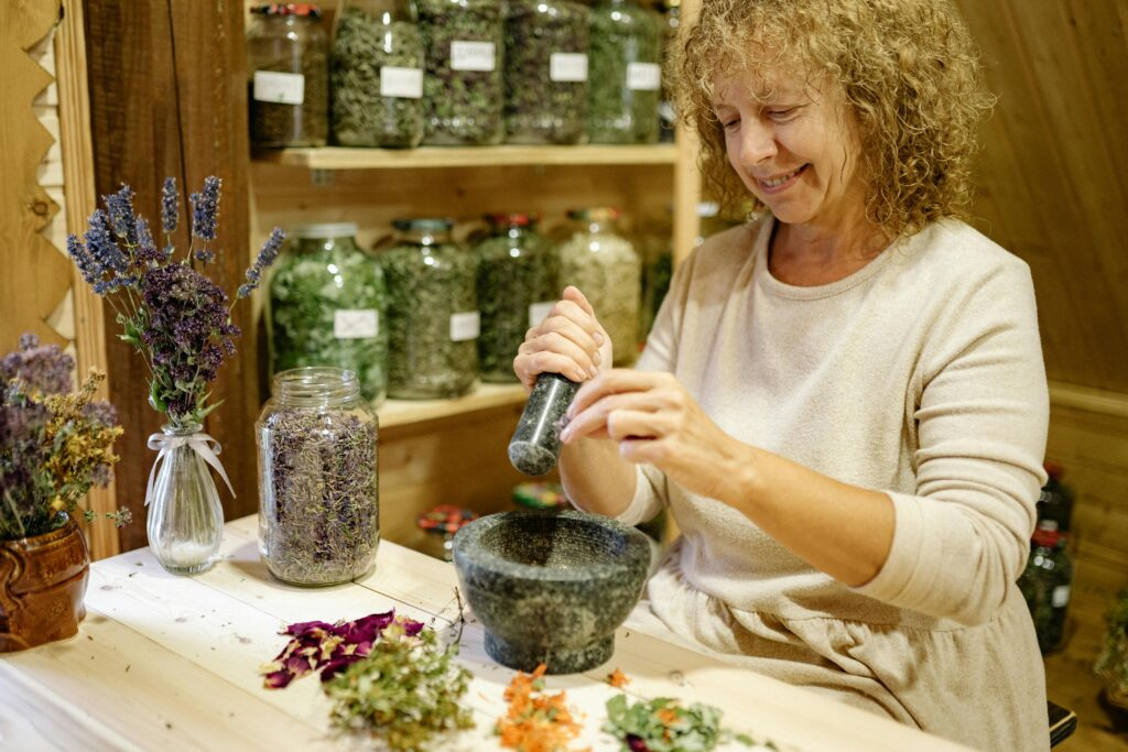 pexels-photo-5480048-5480048 Woman grinding herbs with mortar and pestle in a cozy workshop filled with jars and dried flowers.